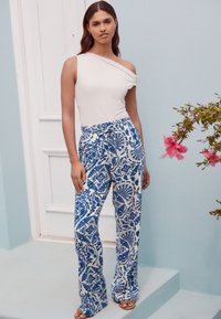 Valkoinen, yhdellä olkapäällä oleva toppi, yhdistetty sinivalkoisiin, leveälahkeisiin housuihin, joissa on kukkakuvioita, vaaleansinisellä taustalla.