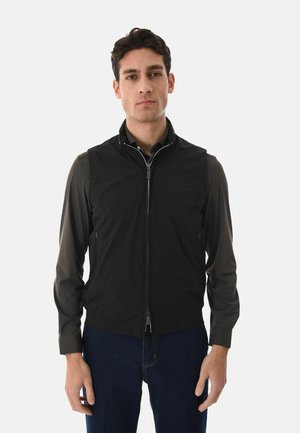 Uomo con indosso un gilet nero con zip sopra una maglia a maniche lunghe scura e jeans blu, in piedi contro uno sfondo bianco semplice.