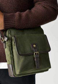 Grüne Canvas-Tasche mit braunen Lederakzenten; verfügt über einen Klappenverschluss, einen Metallschnappverschluss und einen verstellbaren Gurt. Texturiertes Gewebe mit genähten Details.