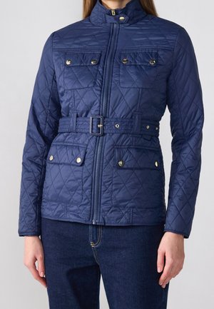 Frau trägt eine dunkelblaue, gesteppte Jacke mit Gürtel und Druckknopftaschen, kombiniert mit dunkelblauen Jeans.