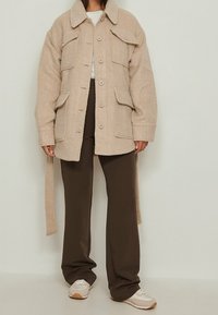 Beige uldblandingsjakke med knaplukning, store brystlommer og spidse kraver. Kombineret med mørkebrune bukser og lyse sneakers.