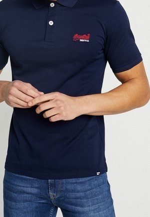 Mężczyzna w granatowej koszulce polo z czerwonym logo na piersi i niebieskich dżinsach, z rękami złożonymi przed sobą na wysokości pasa.