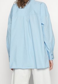 Camicia oversize azzurro chiaro con piega sul retro, polsini abbottonati e texture in cotone morbido. Finestrata con colletto classico e vestibilità comoda.