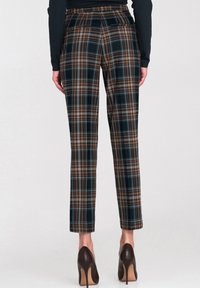 Kobieta w wąskich spodniach w kratę w kolorze czarnym, brązowym i białym, z tylnymi kieszeniami, w połączeniu z czarną bluzką z długim rękawem i ciemnymi szpilkami.