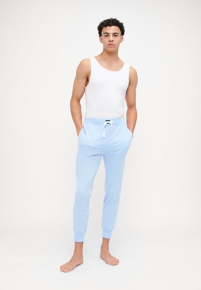 Lyseblå joggebukser med snøring i livet, ribbede mansjetter og sidelommer, kombinert med en hvit tanktop. Bare føtter.