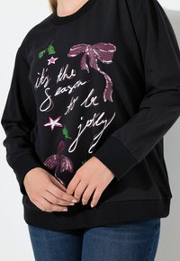 Sudadera negra de manga larga con texto y gráficos de temática navideña en rosa y verde, que incluye un lazo con lentejuelas y acentos de estrellas.