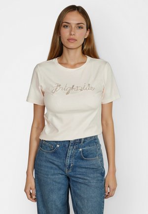 Junge Frau mit langem Haar, die ein weißes T-Shirt mit dem Schriftzug "Brightside" in Strasssteinen und hoch taillierte blaue Jeans trägt, vor weißem Hintergrund steht.