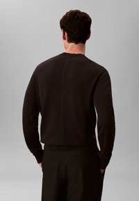 Muž v černém tričku s dlouhým rukávem a černých kalhotách, stojící s rukama v kapsách, otočený zády k šedému pozadí.