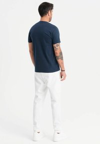 Camiseta de manga corta azul marino combinada con pantalones blancos, con un cuello redondo y tatuajes visibles en los brazos. Calzado informal completa el look.