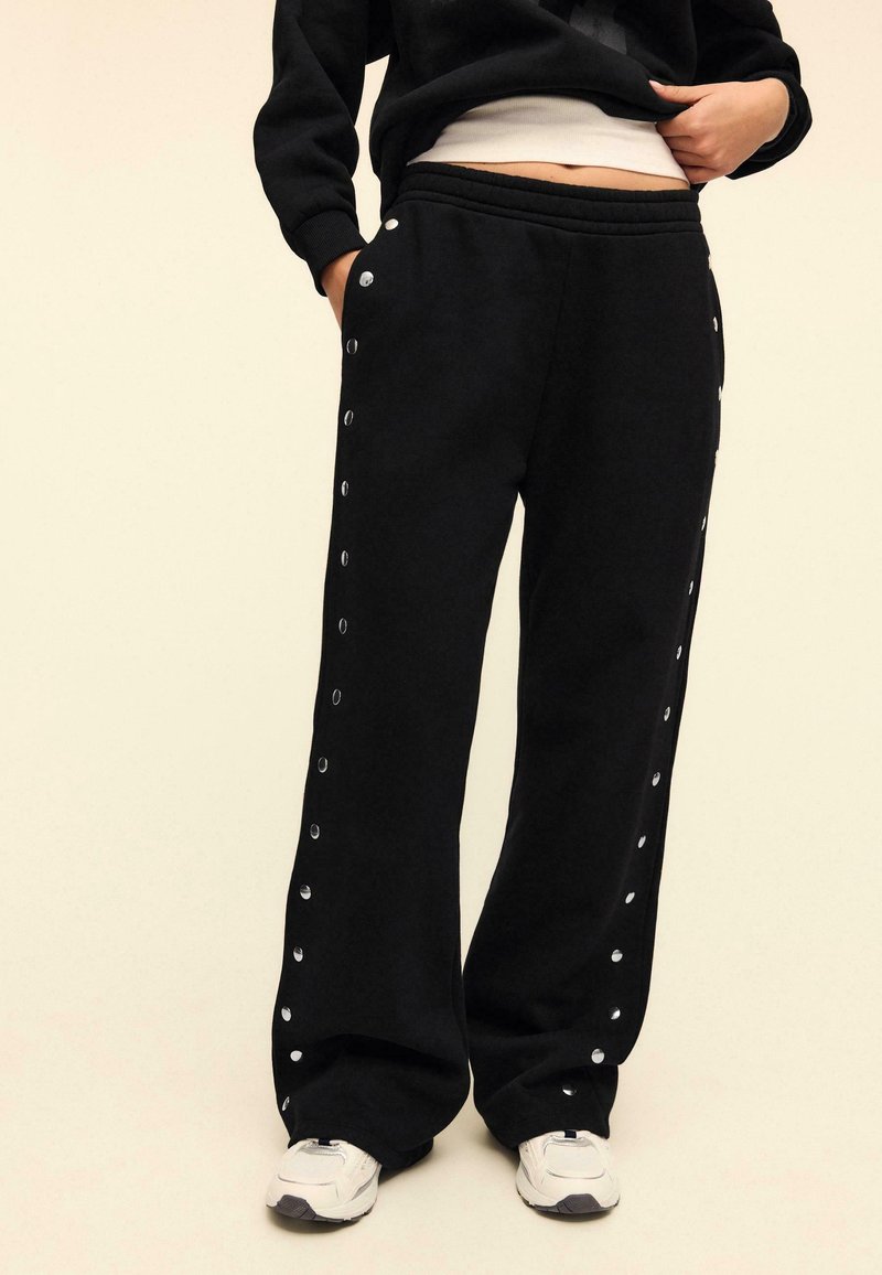 Personne portant un pantalon noir à jambes larges avec des boutons-pression argentés sur les côtés, des baskets blanches et un haut court noir et blanc.