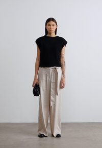 Μαύρο τοπ με κοντά μανίκια, μπεζ παντελόνι σε φαρδιά γραμμή με δεμένη ζώνη και μαύρη τσάντα. Ομαλές υφές και ουδέτερα χρώματα κυριαρχούν στην εμφάνιση.