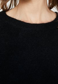 Vrouw in een zwarte donzige trui met een ronde hals, met zichtbare halslijn en gedeeltelijke haarslierten tegen een effen achtergrond.