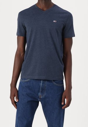 Mann trägt ein eng anliegendes marineblaues V-Ausschnitt-T-Shirt mit einem kleinen Logo auf der Brust und blaue Jeans, steht mit entspannten Armen.