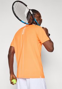 Oransje treningsskjorte med korte ermer, med en vertikal hvit logo og sømmer. Personen holder en tennisracket og ball.