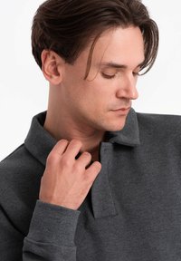 Donkergrijze polo met korte kraag, textuurstof en zichtbare stikseldetails. Model houdt de kraag met één hand vast.