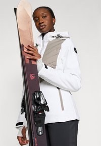 Biela lyžiarska bunda s šedými akcentmi; ľahký materiál. Drží hnedé lyže s čiernymi viazaniami a červeným textom. Čistý, moderný dizajn.