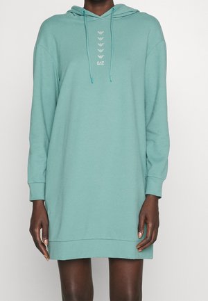Personne portant une robe sweat à capuche longue et ample de couleur vert sarcelle clair avec un logo à l'avant et des cordons, debout sur fond blanc.
