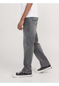 Persona vistiendo jeans rectos grises y zapatillas negras con cordones blancos, caminando contra un fondo liso claro.