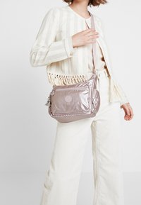 Kobieta w jasnobeżowej, paskowanej marynarce i spodniach trzyma metalicznie różową torbę na ramię z wieloma zamykanymi na zamek kieszeniami.