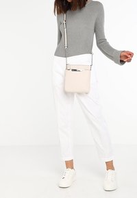 Mujer vistiendo un suéter gris y pantalones blancos, sosteniendo un bolso cruzado beige y con zapatillas blancas, sobre un fondo blanco.