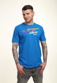 Modré bavlněné tričko s krátkými rukávy, na kterém je barevné grafické logo "JT Racing" umístěné na hrudi. Model má tetování a šedé džíny.