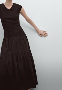 Musta mekko, jossa on hyvin istuva yläosa V-pääntiellä ja hihattomilla hihansuilla, siirtyen kerrokselliseen, valuvaan hameeseen. Liukas puuvillakangas.