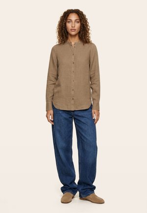 Moteris, stovinti apsirengusi ruda ilgomis rankovėmis palaidine su sagomis, laisvais mėlynais džinsais ir šviesiai rudais bateliais, stovinti priešais vienspalvį foną.