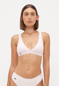 Svetlo roza bralette z V-izrezom, širokimi naramnicami in rebrasto teksturo. Ima majhen zelen logotip na levi strani.