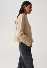 Giacca in pelle beige con colletto, chiusura frontale a bottoni e tasche laterali, abbinata a una gonna in denim scuro. Texture liscia, design corto.