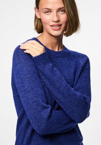 Karaliski zilā adīta džemperis ar garām piedurknēm un ribotām aprocēm, ar klasisku apaļu kakla izgriezumu un mīkstu, teksturētu apdari.