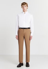 Junger Mann steht, trägt ein weißes Hemd mit Knöpfen, beige Hose mit schwarzem Gürtel und dunkelbraune Anzugschuhe, vor einem schlichten Hintergrund.
