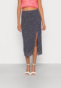 Jupe midi imprimée florale marine avec une fente latérale, tissu léger et design fluide, associée à des sandales blanches à talons.