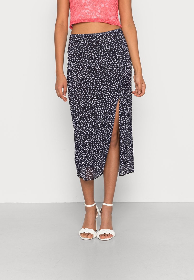 Jupe midi imprimée florale marine avec une fente latérale, tissu léger et design fluide, associée à des sandales blanches à talons.