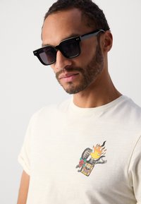 Hvid bomulds t-shirt med et farverigt grafisk motiv, der viser et kranium, flammer og en rose. Sorte rektangulære solbriller med mørke linser.