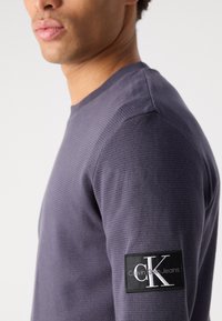 Lila waffle kötésű, hosszú ujjú póló, kerek nyakkivágással és a bal ujján "Calvin Klein Jeans" logóval ellátott márkás folttal.
