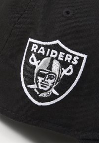 Gorra negra con un logo de los Raiders bordado en blanco, que incluye una forma de escudo, una figura con casco y espadas cruzadas.