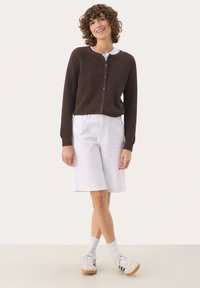 Brauner, gerippter Strick-Cardigan mit Knopfleiste, kombiniert mit weißen, knielangen Jeansshorts und weißen Sneakers mit schwarzen Streifen.