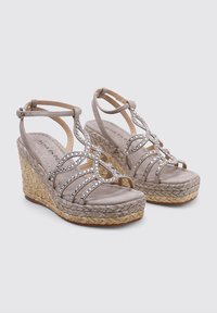 Sandalias de cuña beige con una base de yute trenzado. Presentan correas metálicas adornadas con strass transparentes. Plantilla suave y texturizada con correas ajustables en el tobillo.