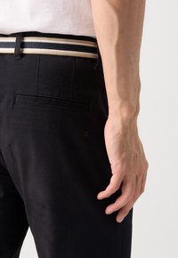 Black trousers with a smooth texture, featuring a back pocket, paired with a multicolored striped belt. Hand resting on the hip.