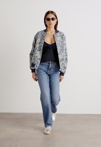 Blauwe bloemen bomberjack, zwarte top, lichtblauwe spijkerbroek en zilveren sneakers. Model draagt zwarte zonnebril en staat op een neutrale achtergrond.