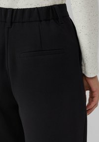 Czarne spodnie o gładkiej teksturze, z elastycznym pasem i dyskretną tylną kieszenią, prezentowane obok jasnego topu w cętki.