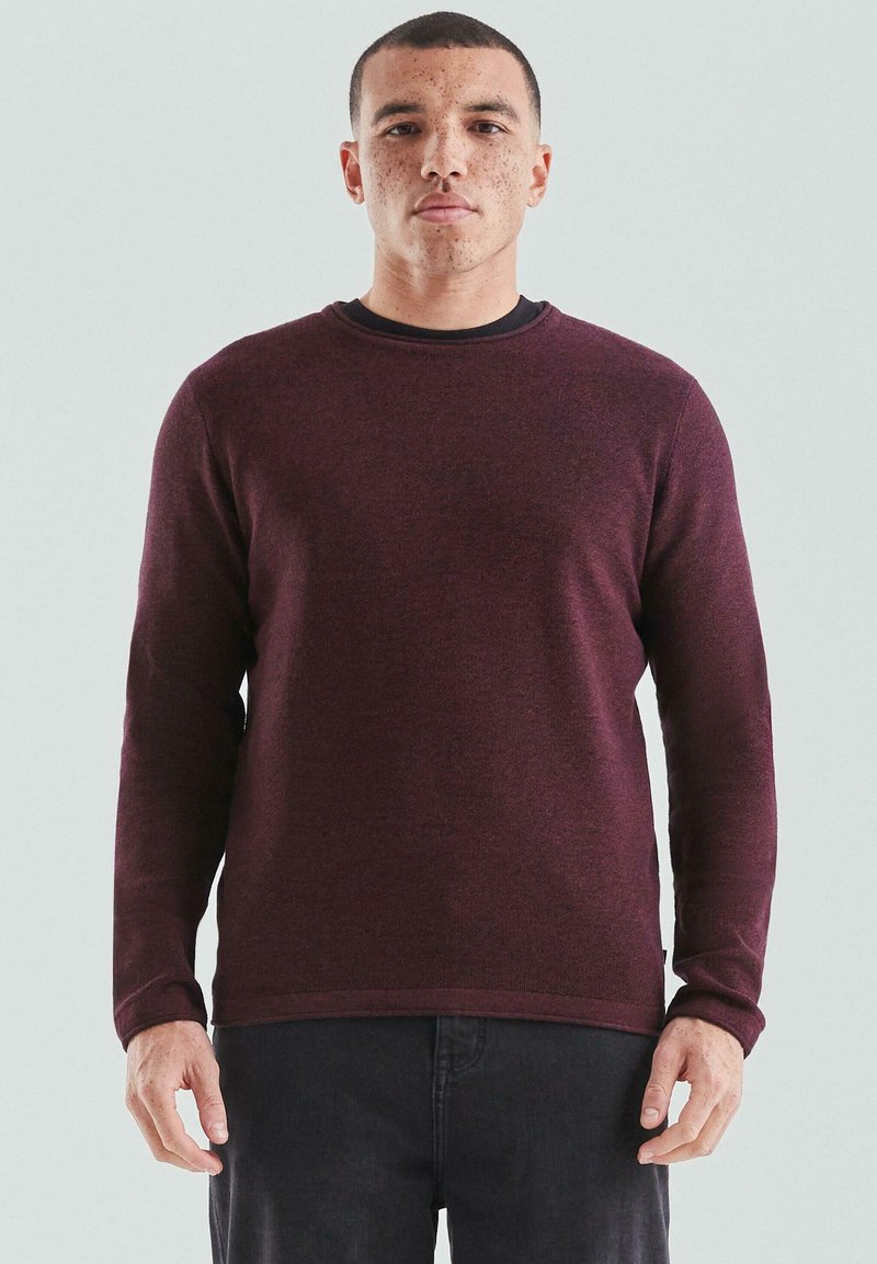 Mężczyzna z krótkimi włosami i piegami, ubrany w długi rękaw ciemnoczerwonego swetra typu crewneck i czarne spodnie, na jednolitym jasnym tle.