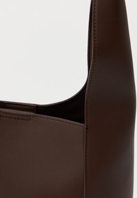 Hnedá kožená kabelka cez rameno s hladkým povrchom, zakriveným tvarom a minimálnym prešívaním. Disponuje vrchným otvorom bez viditeľného kovania.