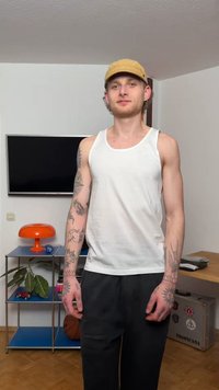 Junger Mann mit Tätowierungen an Armen und Händen, trägt eine beige Mütze, ein weißes Tanktop und schwarze Hosen, steht in einem modernen Wohnzimmer.