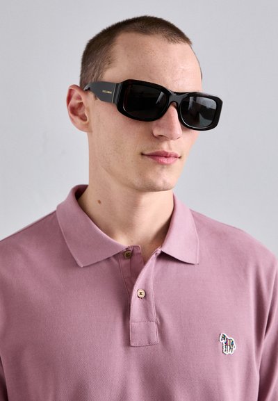 Svarte rektangulære solbriller med mørke linser og gullbokstaver, båret av en person i en lys rosa polo-skjorte med et lite brodert emblem.