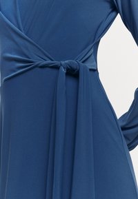 Robe enveloppante bleue en tissu lisse avec un corsage ajusté et un lien à la taille, dotée de manches longues et de détails drapés.