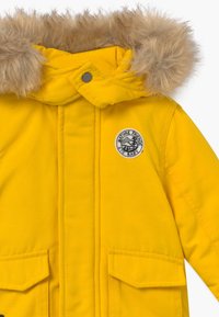 Veste jaune isolante avec une capuche doublée de fausse fourrure, deux poches avant, et un patch logo circulaire arborant un motif de montagne.
