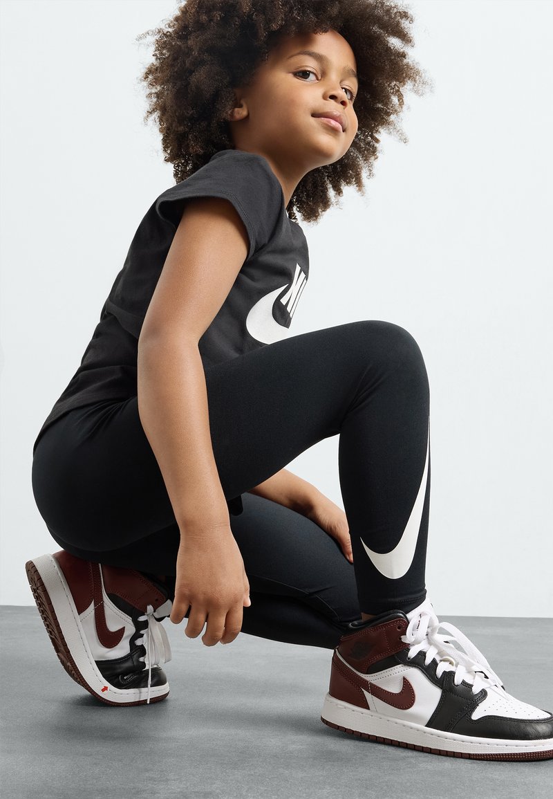 Fekete Nike póló nagy logóval, fekete leggings az oldalsó logóval és barna/fehér magasszárú cipők. Texturált haj, guggoló póz.