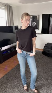 Persona joven con pelo rubio que lleva una camiseta negra, vaqueros azul claro y zapatos marrones, de pie en un salón con televisor y puf.