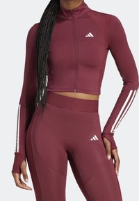 Kvinna iklädd en vinröd långärmad kort zip-up topp och matchande högmidjade leggings med vita Adidas-logotyper och ränder på ärmarna.